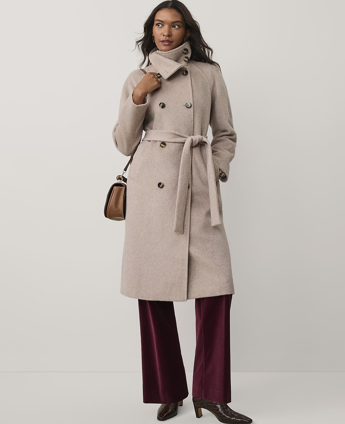 A woman wearing a beige belted coat and burgundy velvet trousers stands against a plain white background.