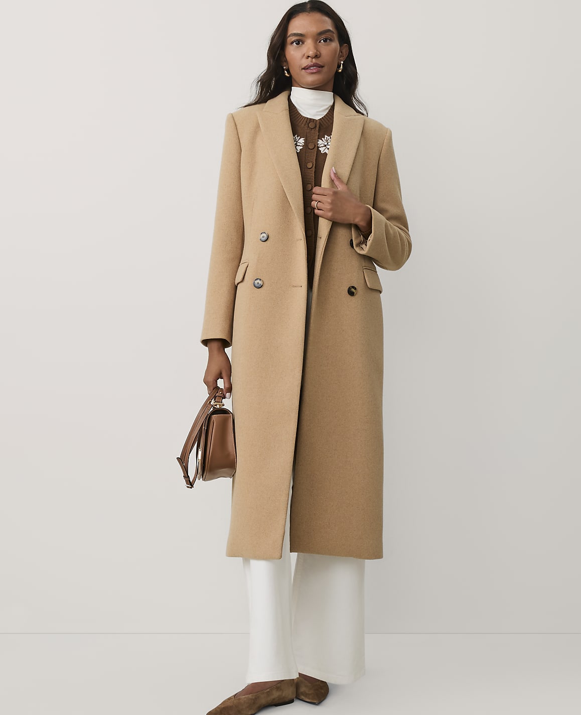 A woman wearing a long camel-colored coat, white trousers, and brown shoes stands against a plain white background.