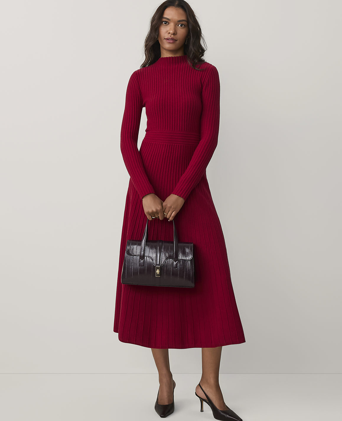 A woman wearing a long-sleeved red ribbed dress and black heels holds a dark burgundy handbag against a plain white background.