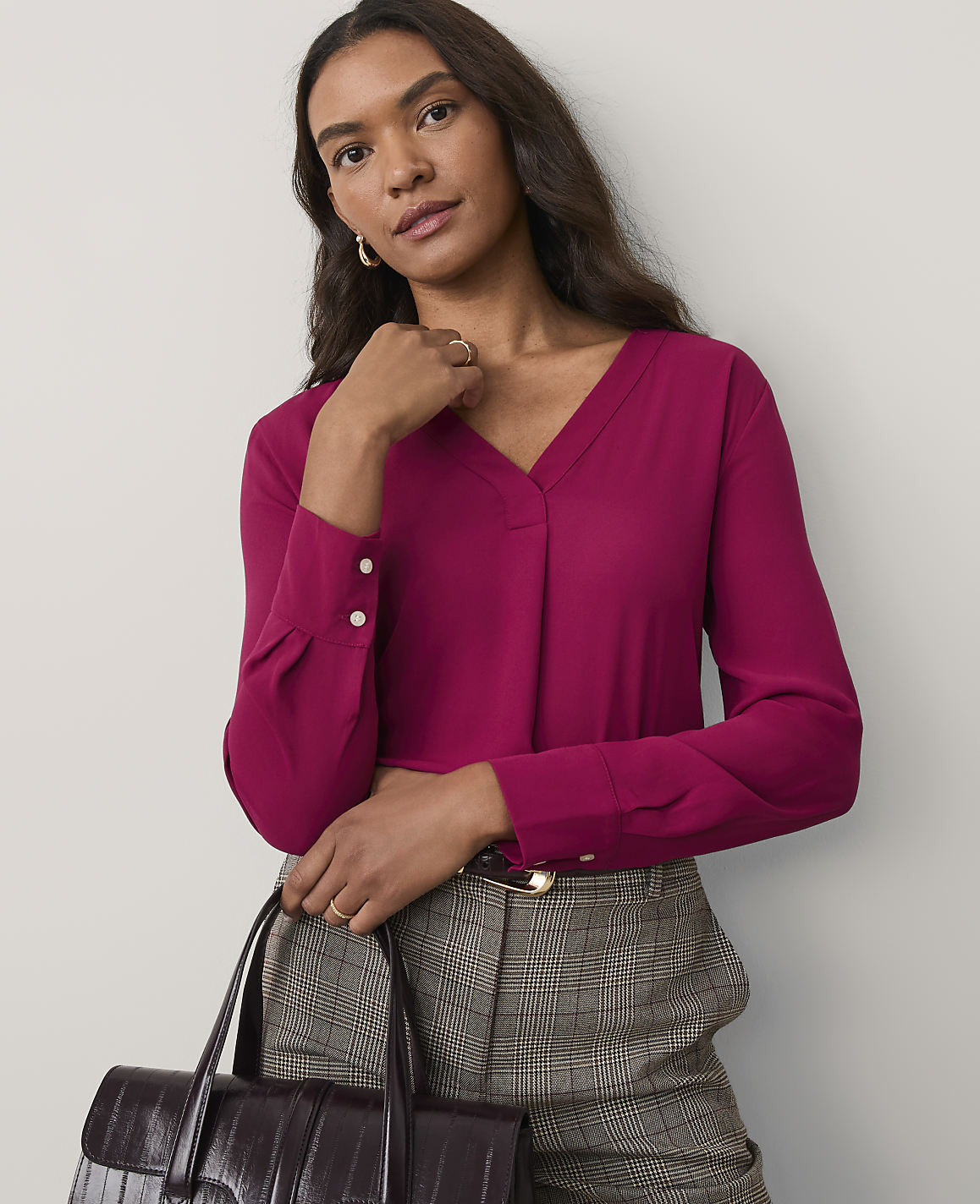 A woman wearing a magenta v-neck blouse and plaid trousers stands against a plain, light-colored wall.