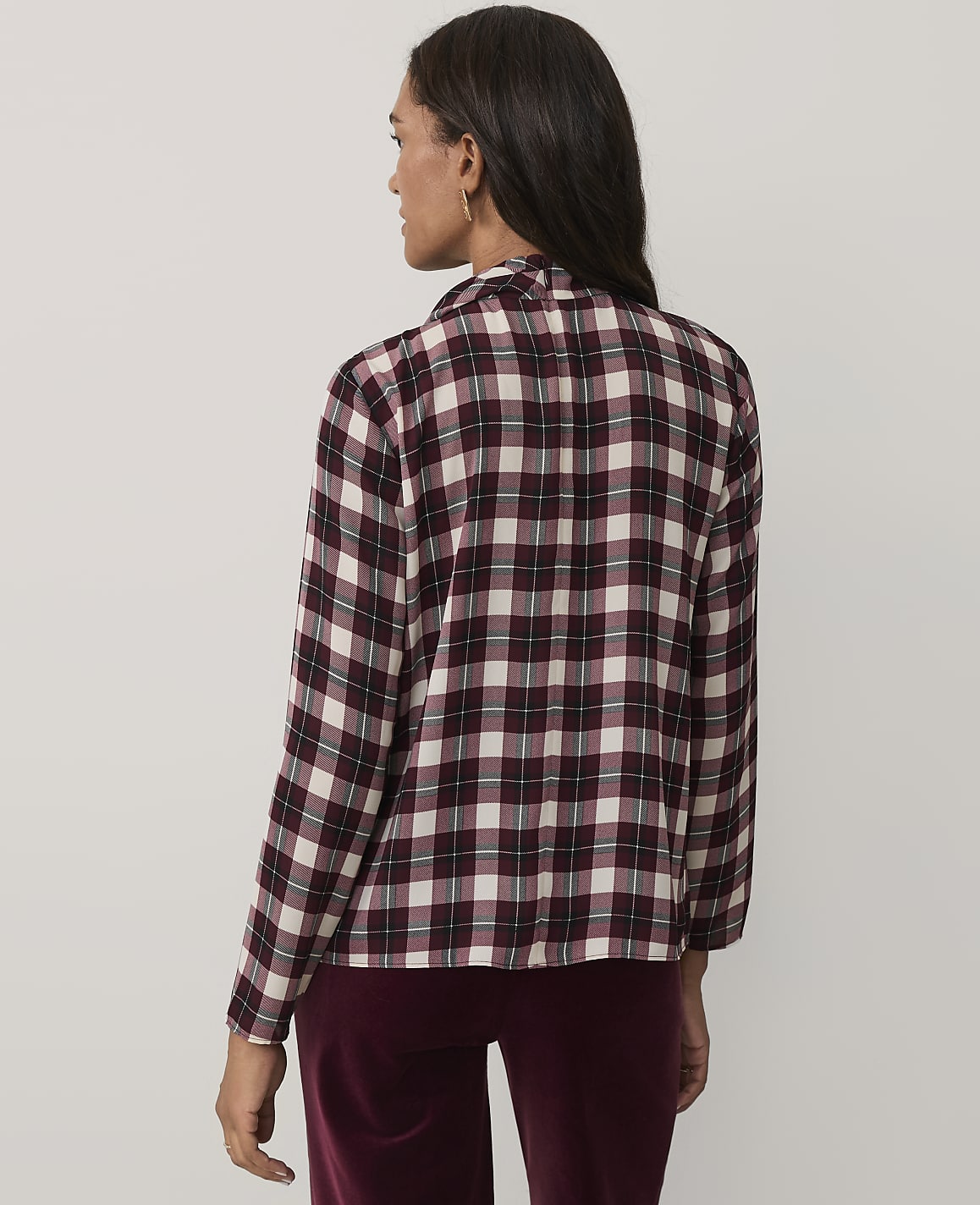 A woman wearing a maroon and white plaid shirt and maroon velvet pants stands against a plain light gray background.
