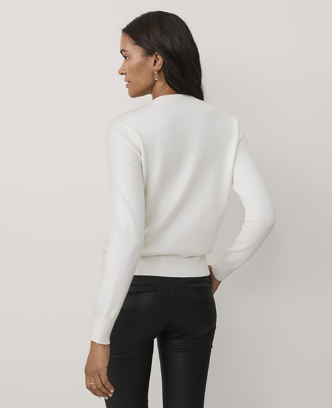 A woman wearing a white sweater and black leather pants stands against a plain, light-colored background.