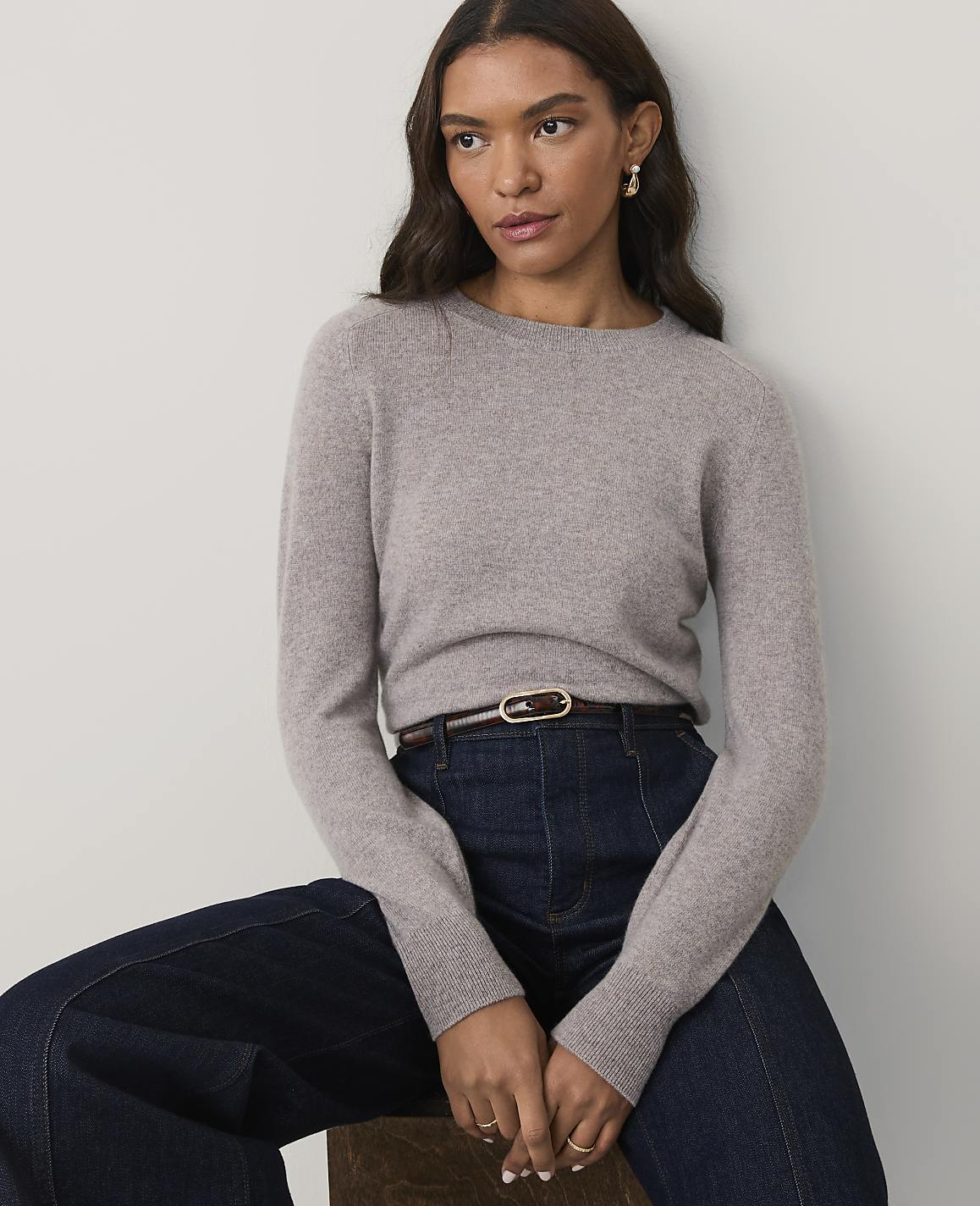 A woman wearing a grey sweater and dark blue jeans sits against a plain white background.