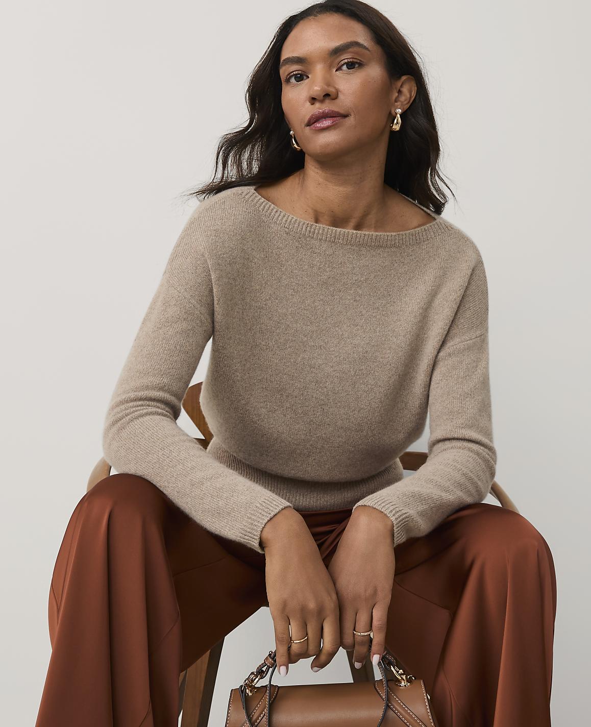 A woman wearing a beige sweater and rust-colored satin pants sits in a wooden chair against a plain white background.