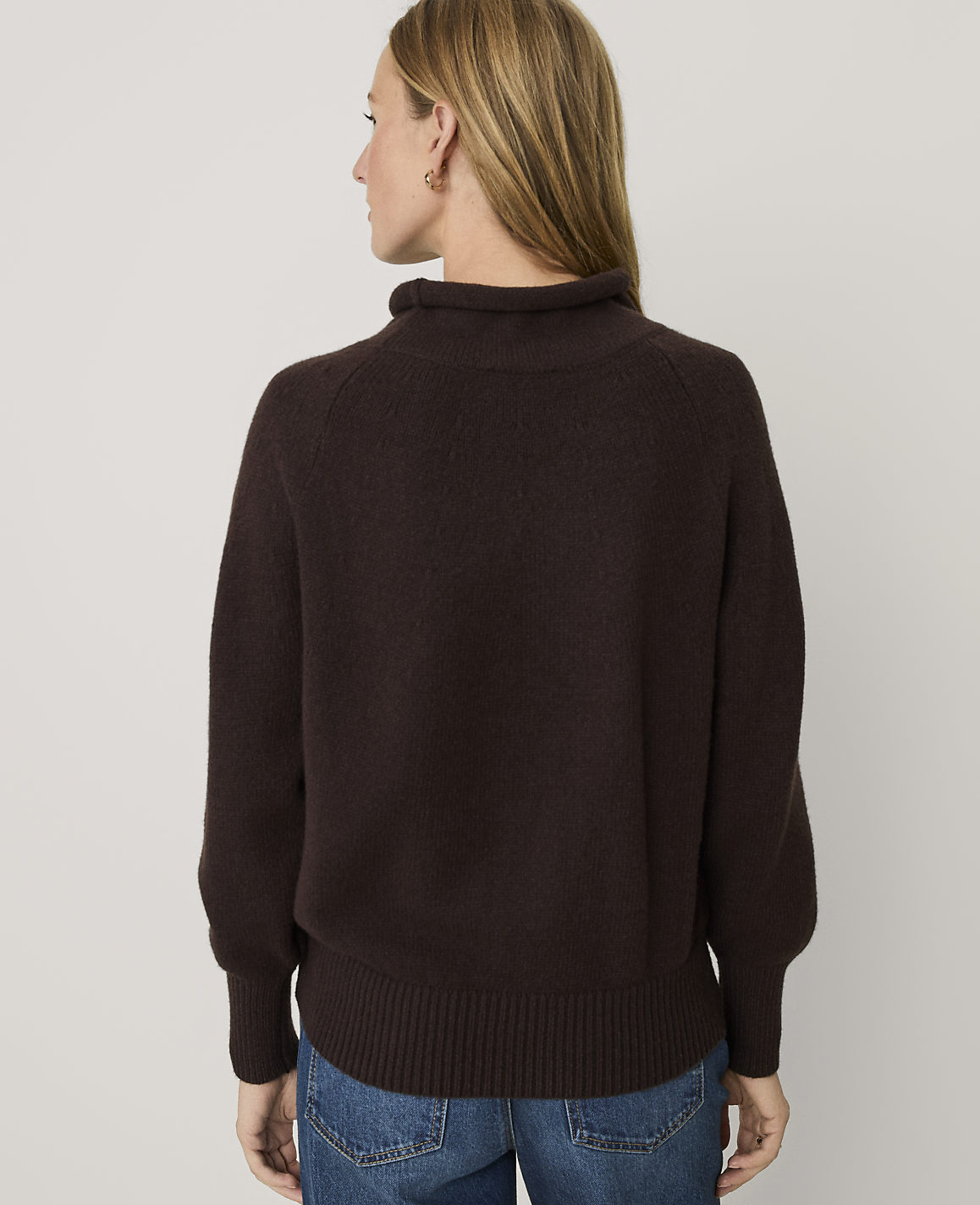 A woman wearing a dark brown knit sweater and blue jeans stands with her back to the camera against a plain white background.