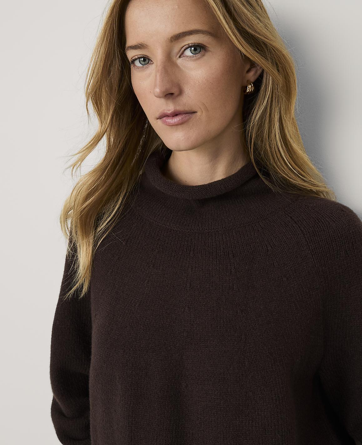 A blonde woman wearing a dark brown turtleneck sweater and blue jeans sits against a plain, light-colored background.