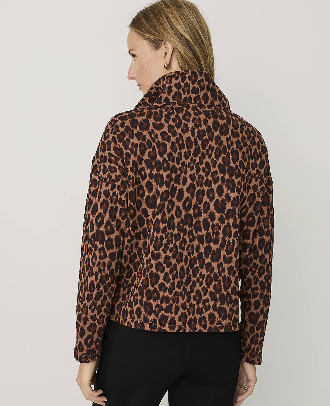 A woman wearing a leopard-print jacket and black pants stands against a plain light gray background.