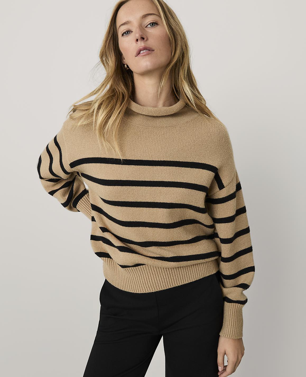 A woman wearing a tan and black striped sweater and black pants stands against a plain light gray background.