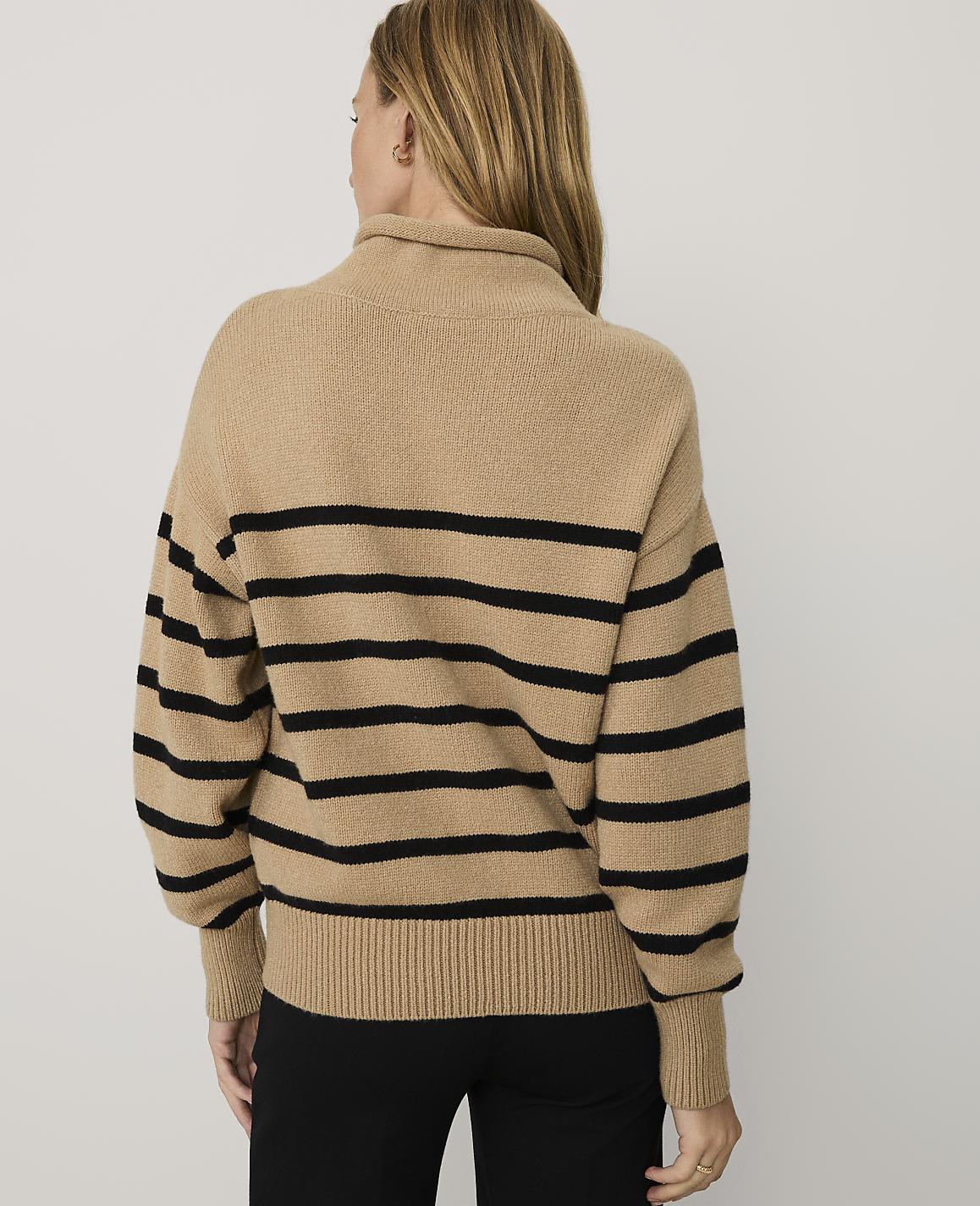 A woman wearing a tan and black striped sweater and black pants stands against a plain white background.