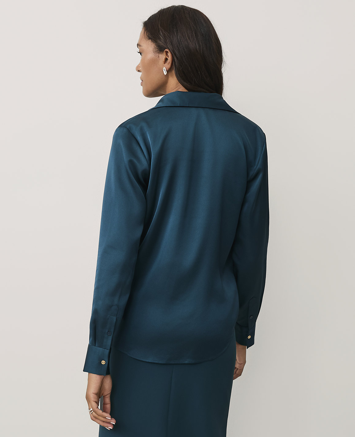 A woman wearing a teal satin shirt and matching skirt stands with her back to the camera against a plain light gray background.