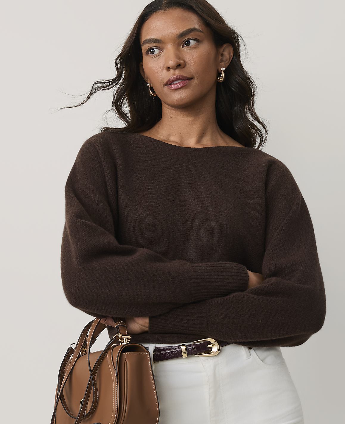 A woman wearing a brown sweater and white pants stands with her arms crossed against a plain light gray background.
