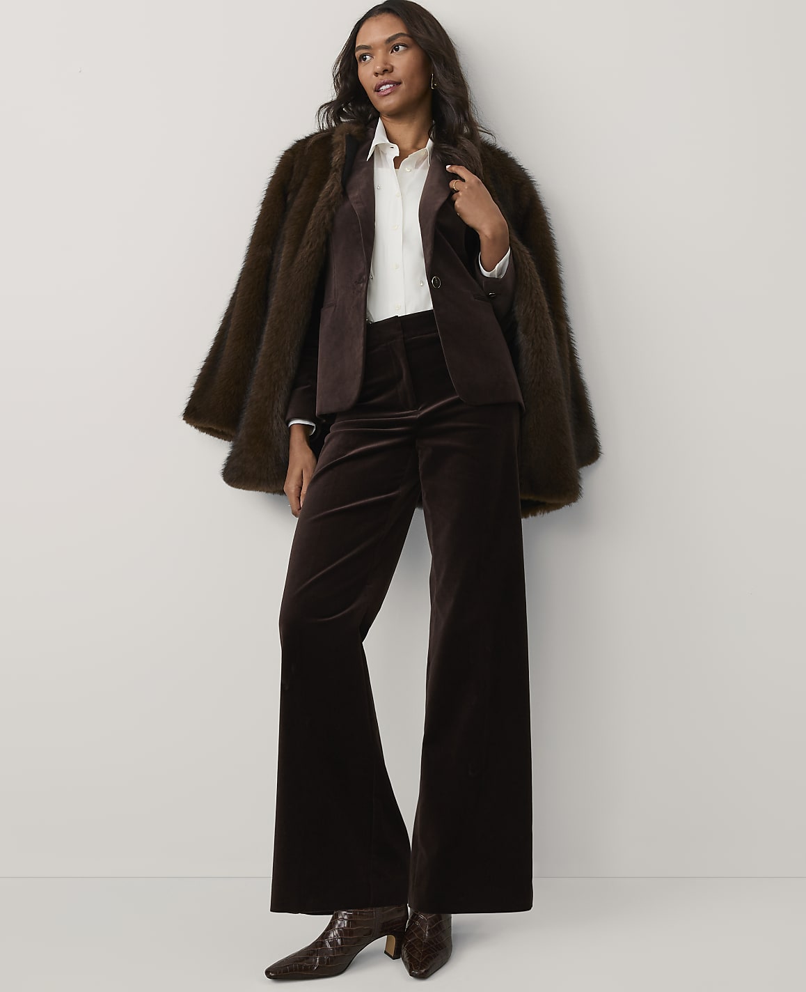 A woman wearing a brown faux-fur coat, blazer, and wide-leg velvet trousers stands against a plain white background.