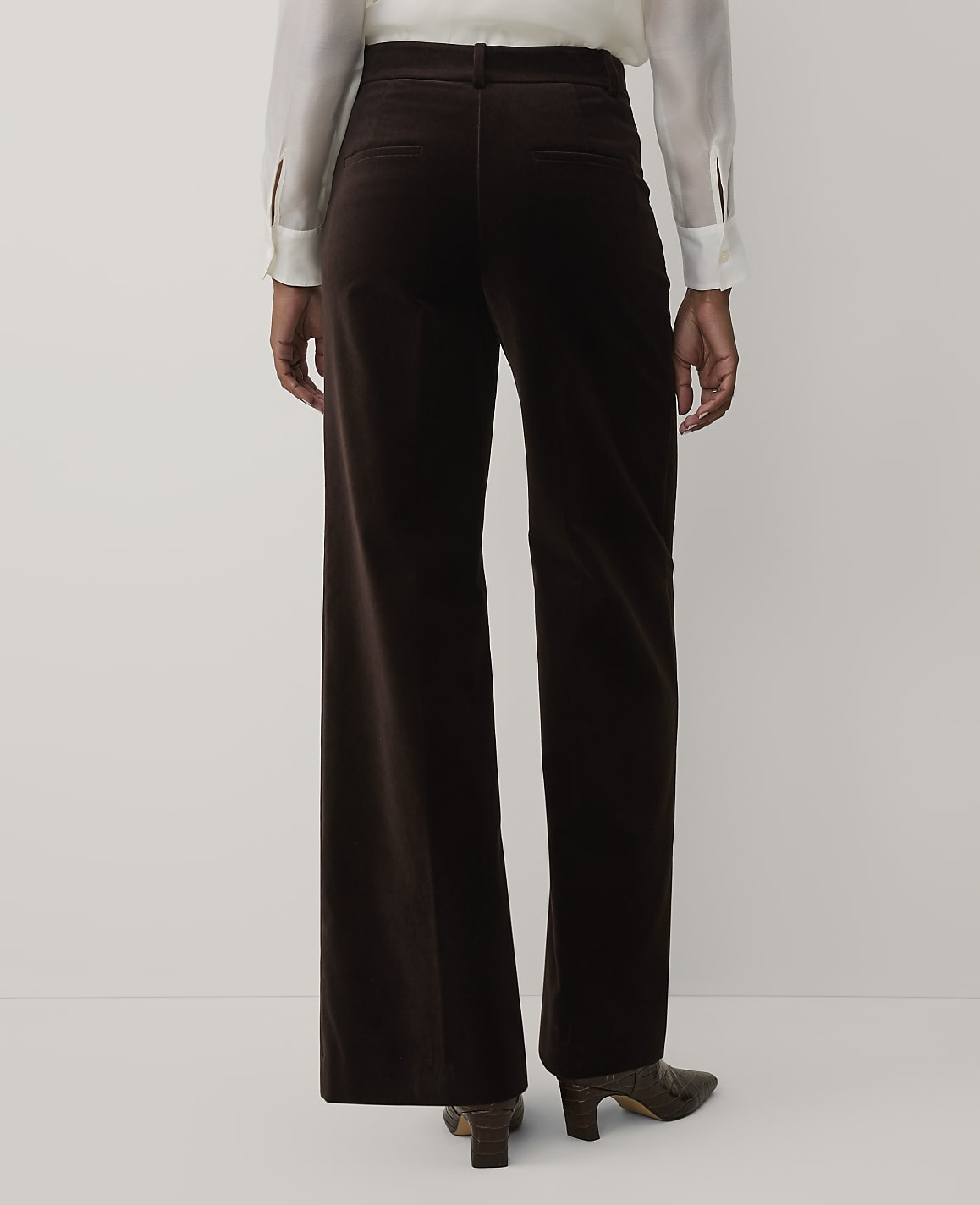A person wearing a white blouse and dark brown velvet wide-leg trousers stands against a plain light gray background.