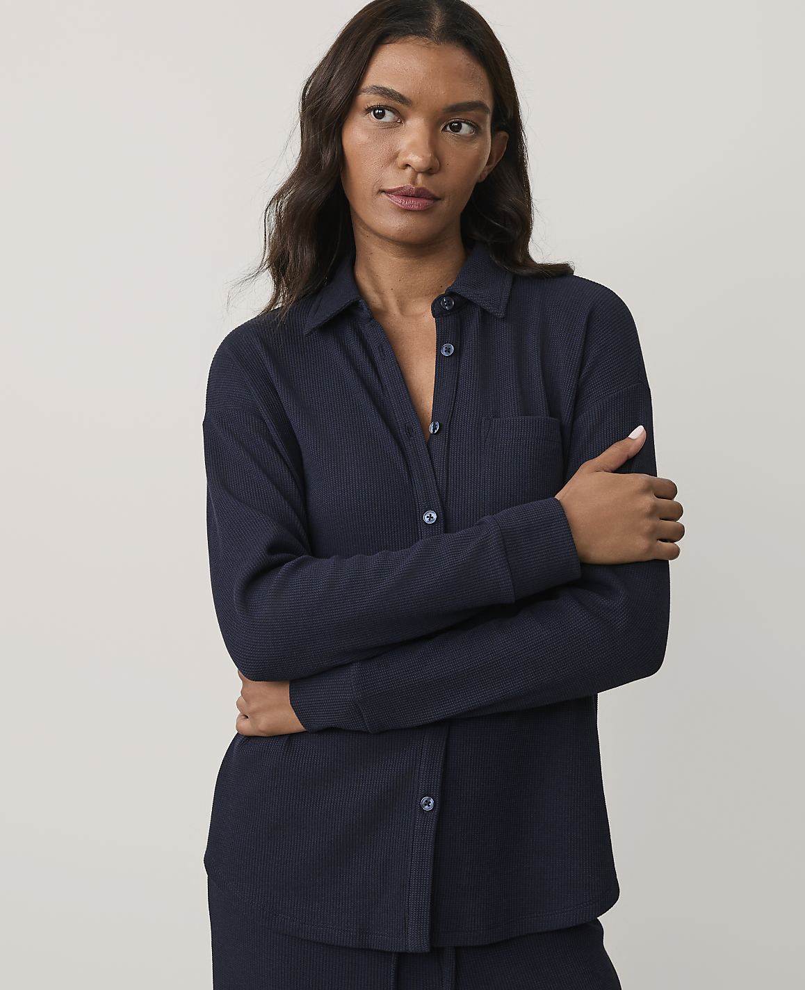 A woman wearing a navy blue ribbed button-down shirt stands with her arms crossed against a plain, light grey background.