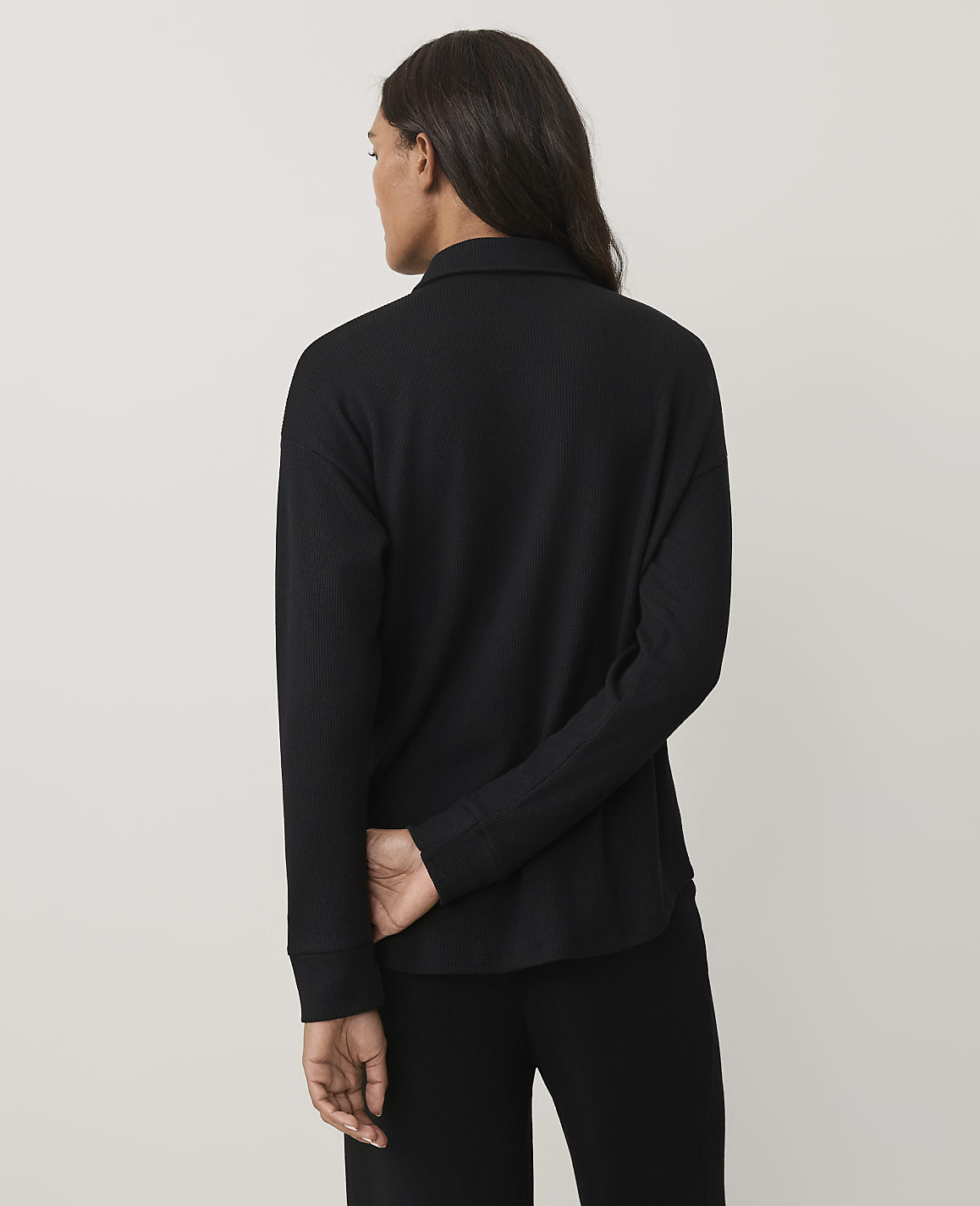 A woman wearing a black long-sleeved ribbed top and black pants stands with her back to the camera against a plain light gray background.