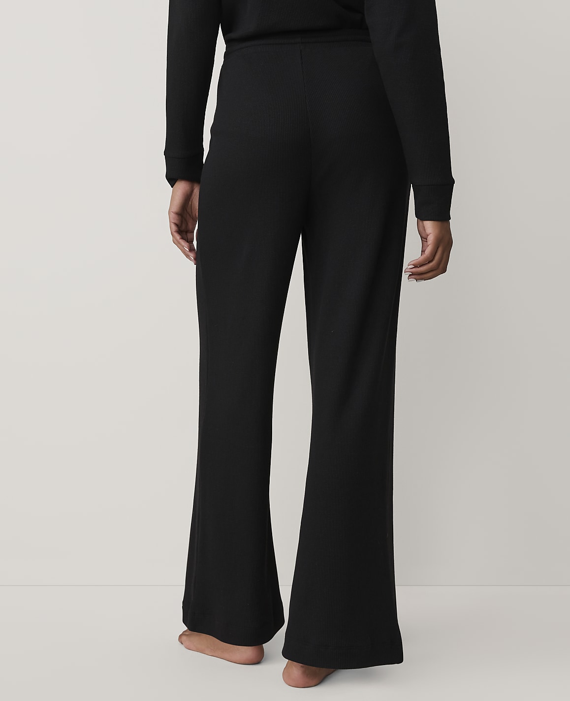 A person stands with their back to the camera wearing black ribbed wide-leg pants and a matching top against a plain light grey background.
