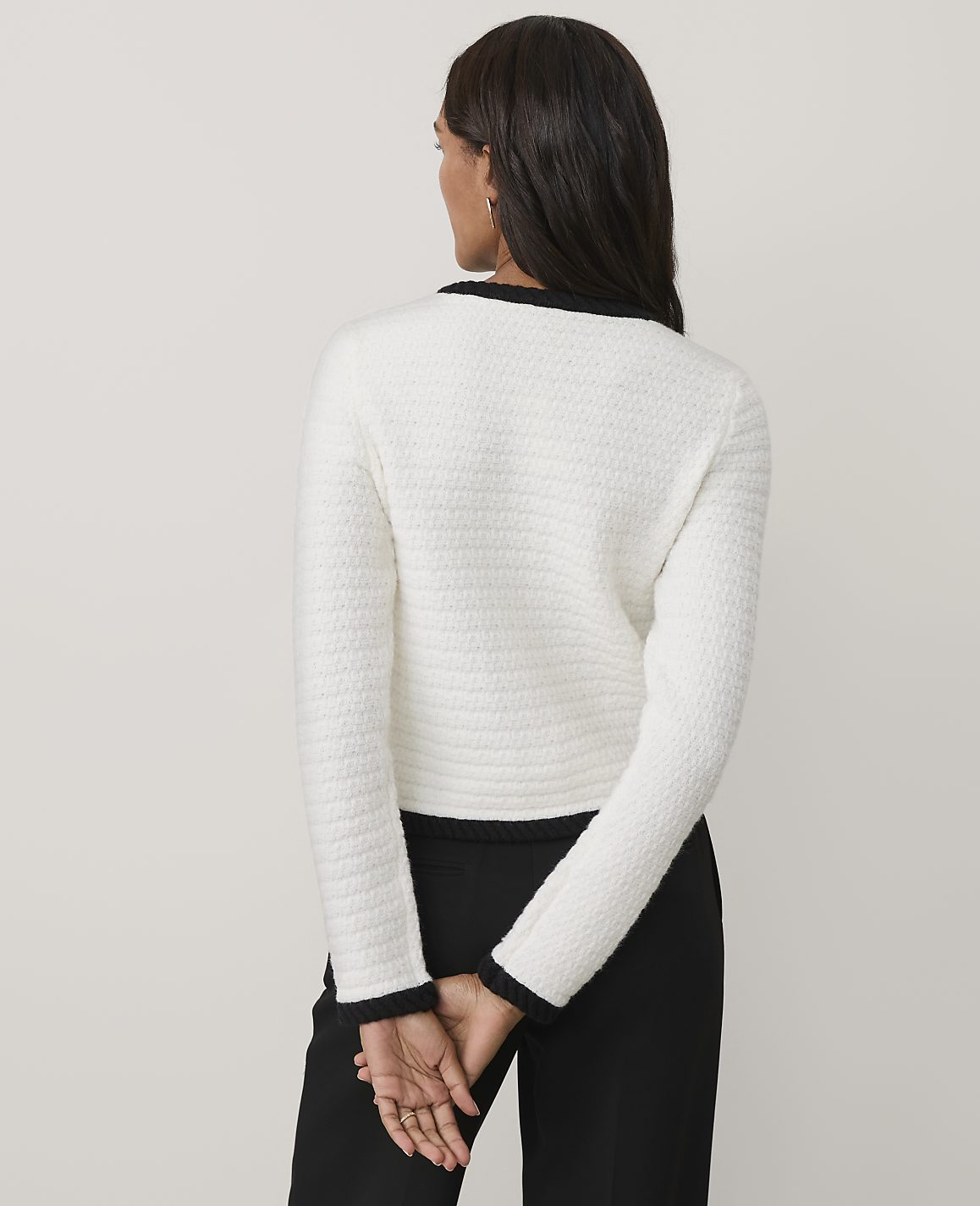 A woman wearing a white textured jacket and black trousers stands with her back to the camera against a plain, light-colored background.
