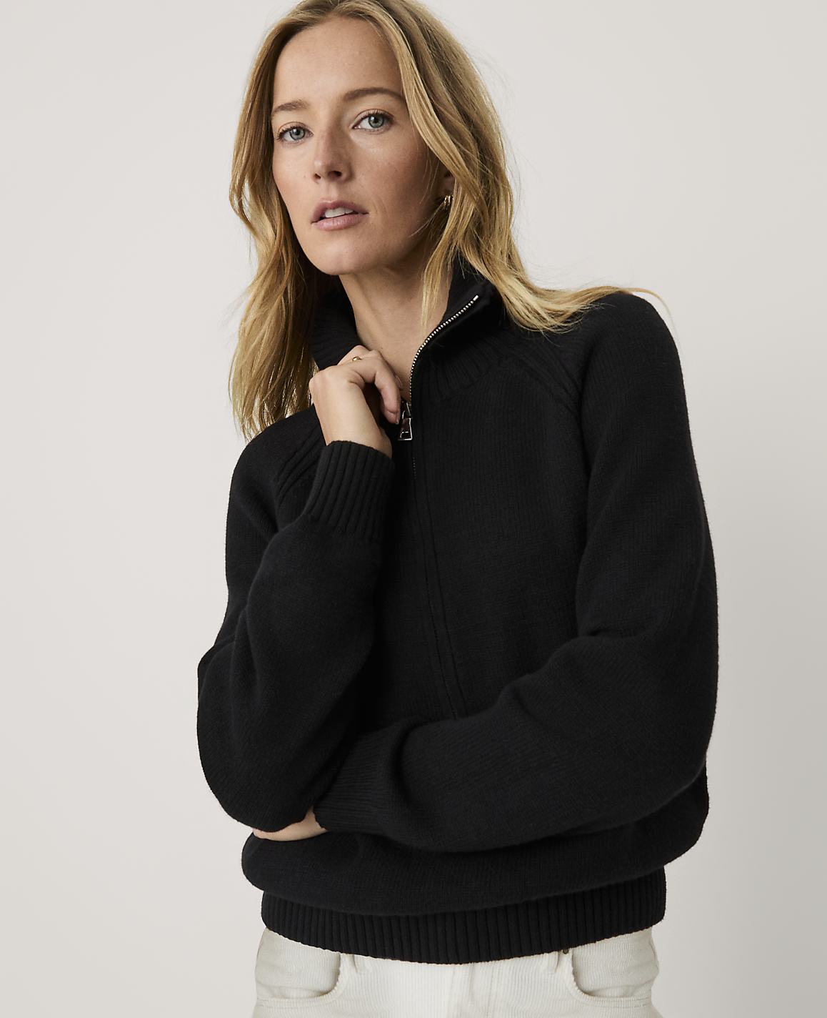 A blonde woman wearing a black zip-up sweater and white pants stands against a plain, light-colored background.