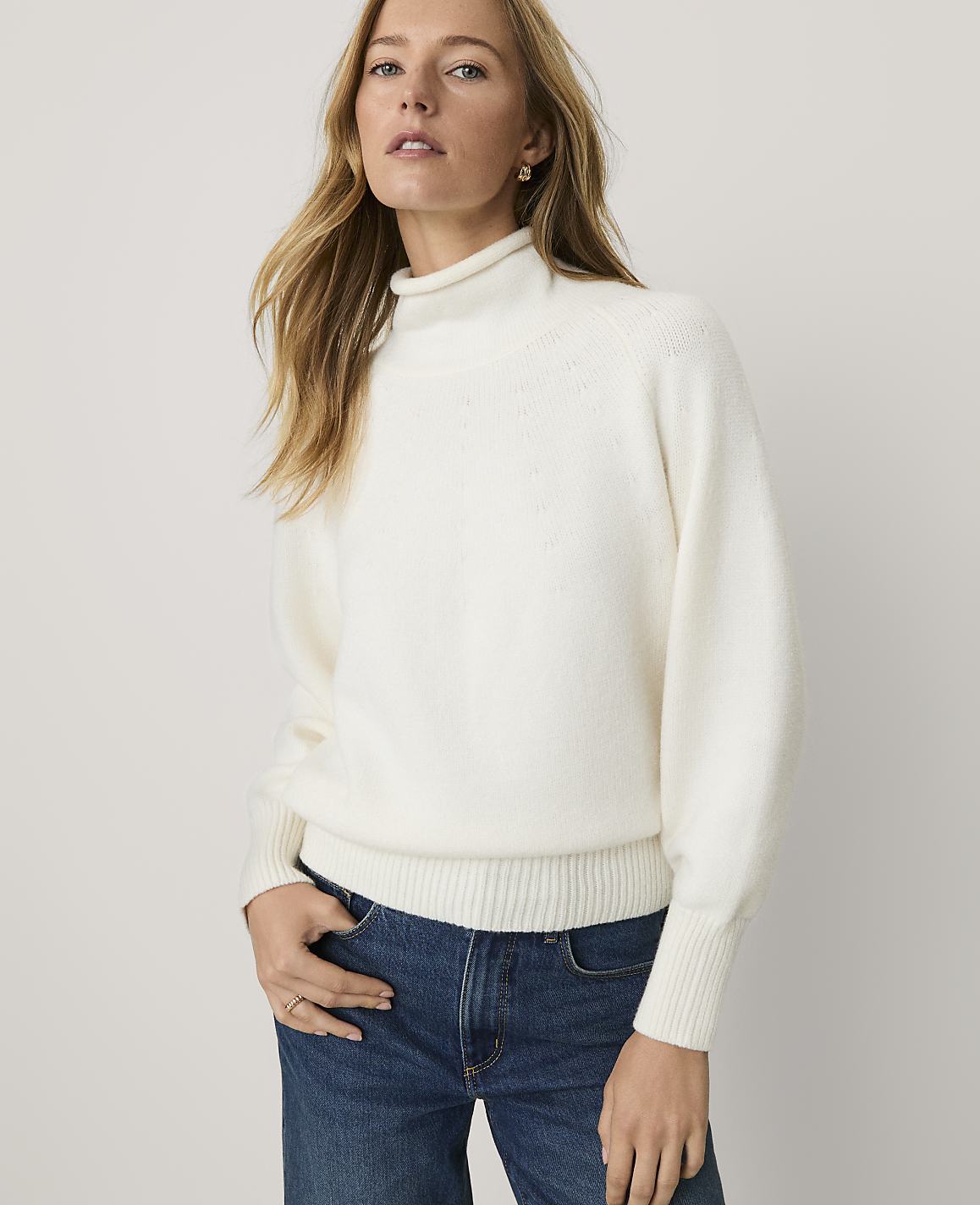 A woman wearing a white turtleneck sweater and blue jeans stands against a plain, light-colored background.