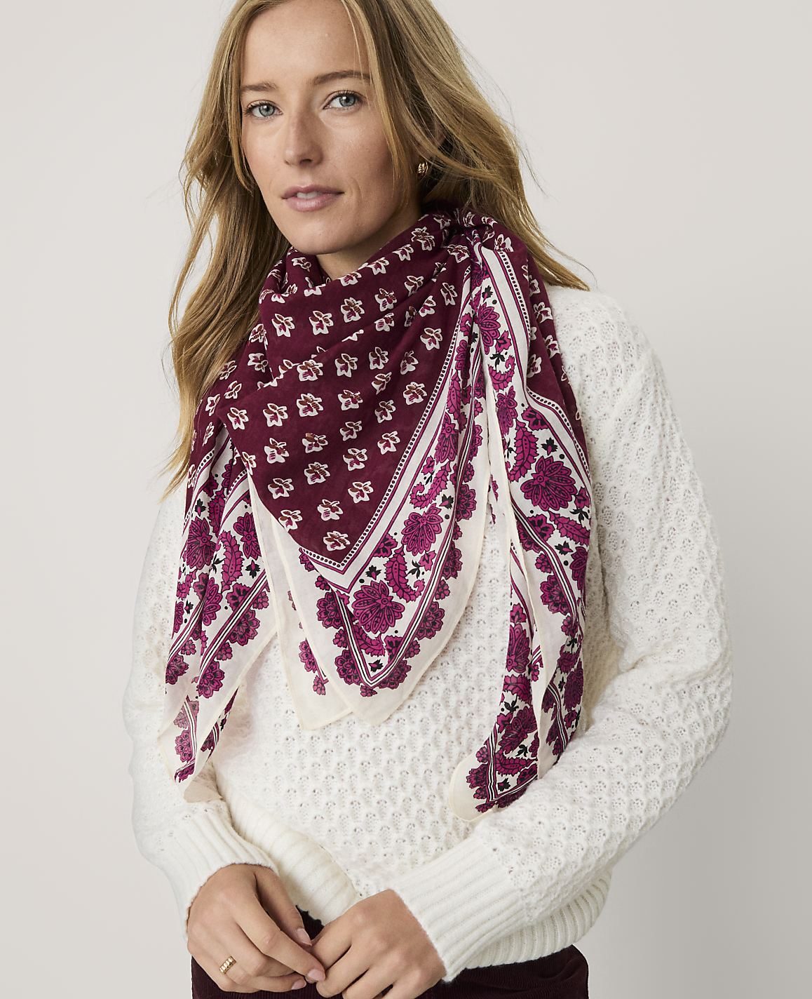 A woman wearing a white textured sweater and a maroon patterned scarf stands against a plain light-colored background.