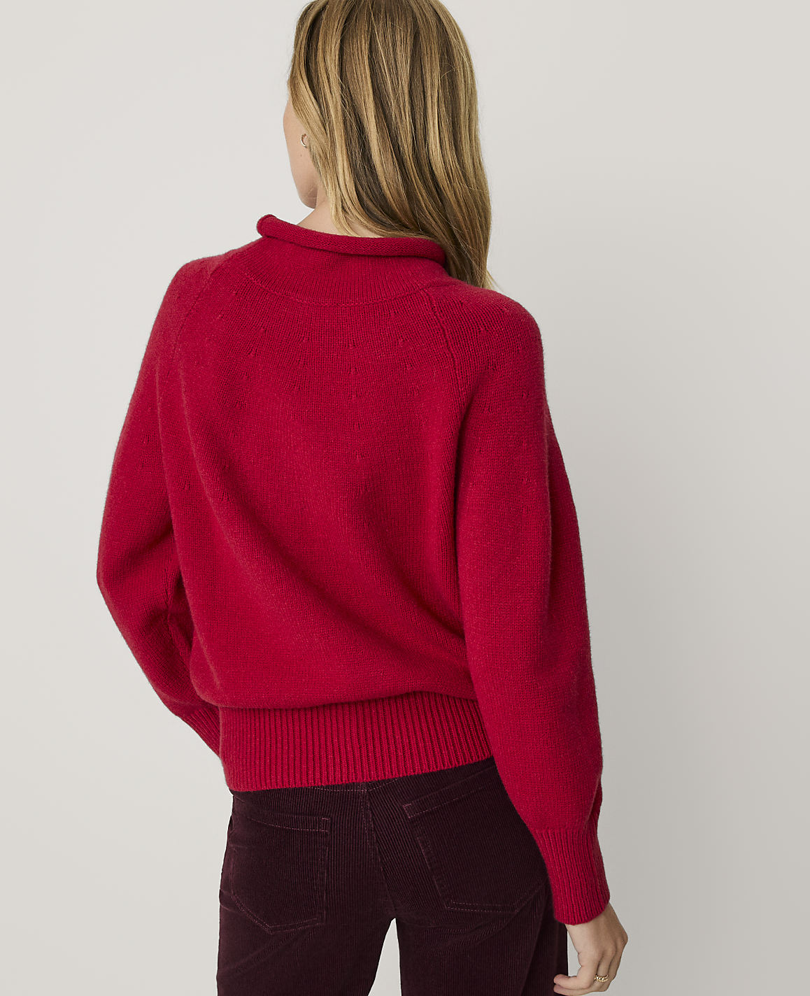 A woman wearing a bright red sweater and dark burgundy corduroy pants stands against a plain white background.