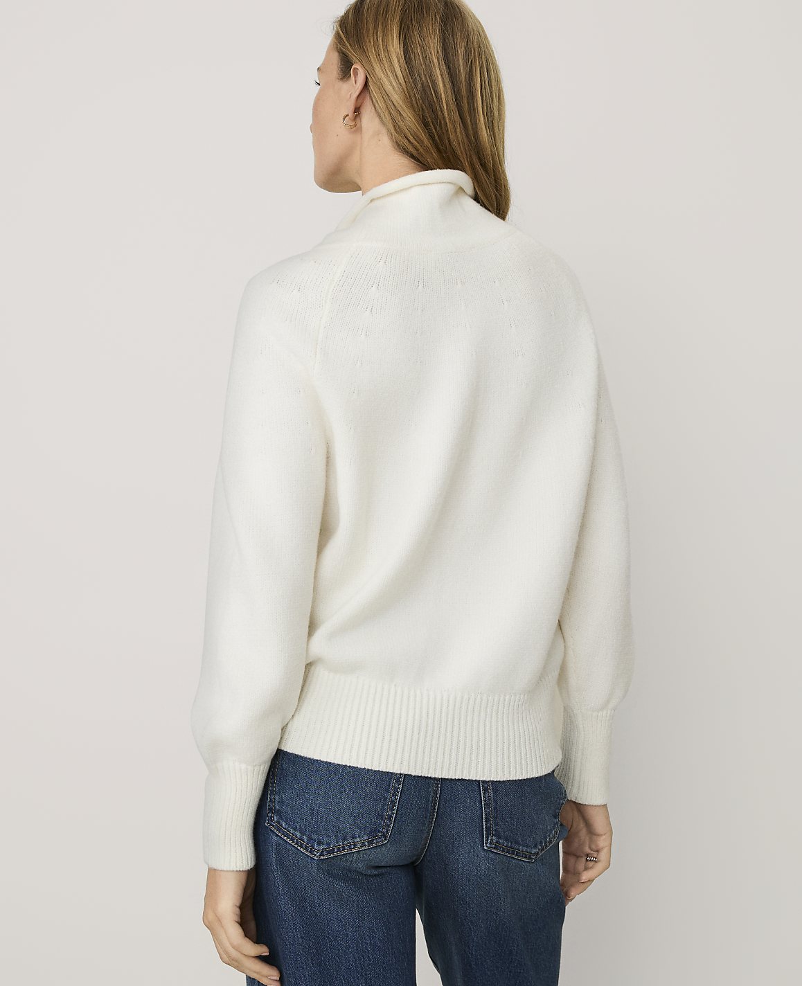 A woman wearing a white fuzzy sweater and blue jeans stands with her back to the camera against a plain light gray background.