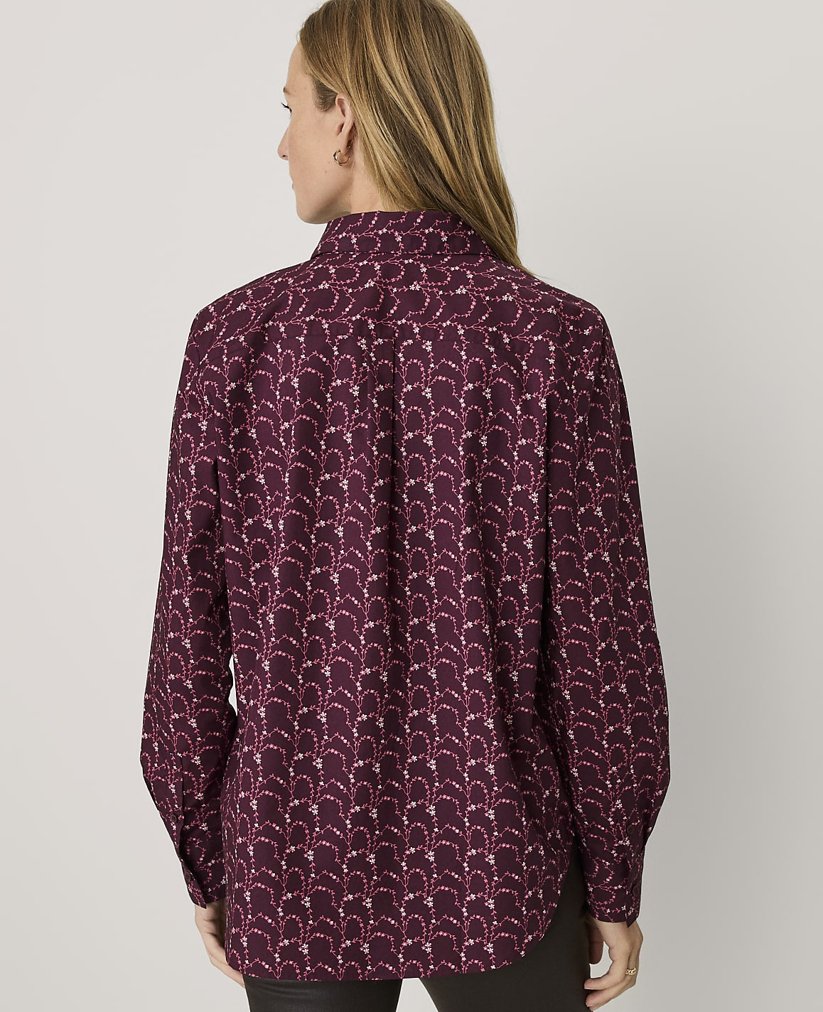 A woman with long blonde hair is seen from behind wearing a maroon patterned button-down shirt against a plain light gray background.