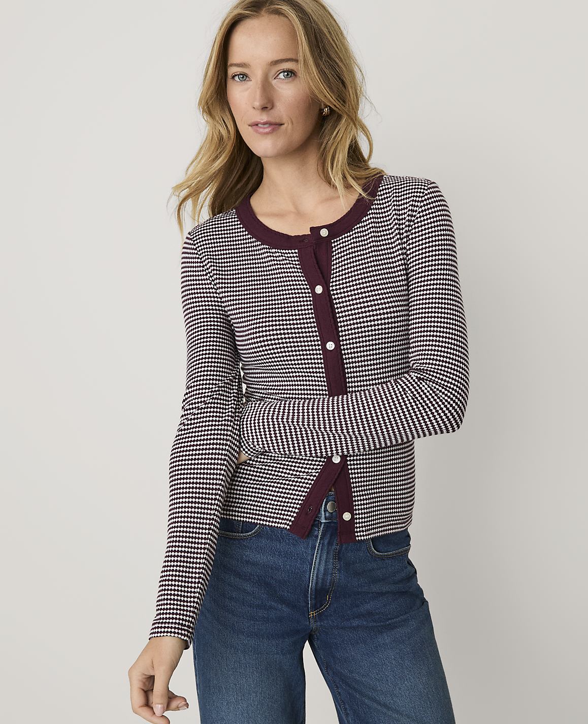 A blonde woman wearing a maroon and white checkered cardigan and blue jeans stands against a plain light gray background.