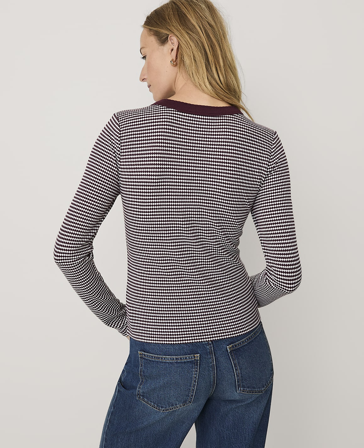 A woman wearing a maroon and white checkered long-sleeve shirt and blue jeans stands against a plain white background.