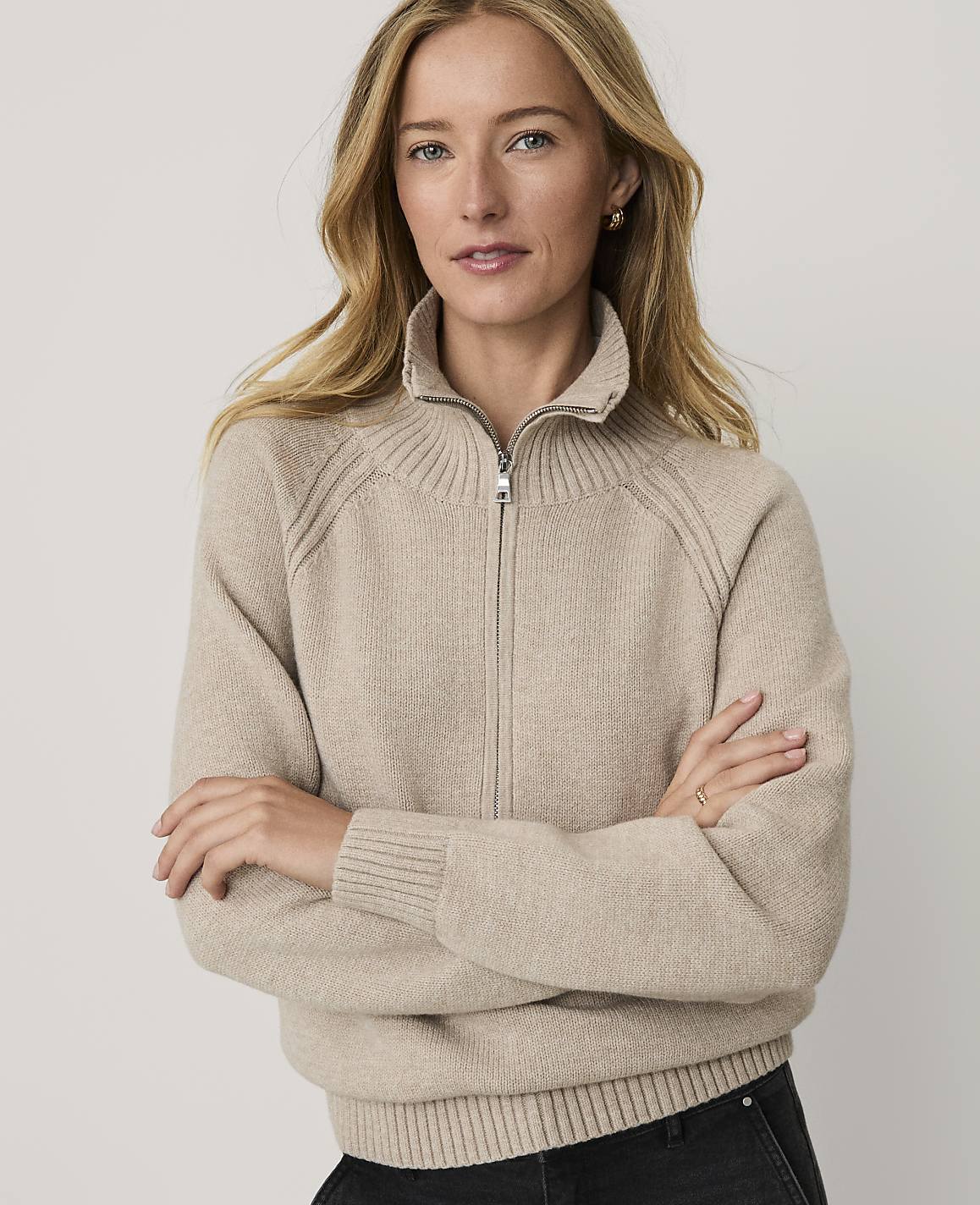 A woman with blonde hair wearing a beige zip-up sweater stands with her arms crossed against a plain, light-colored background.