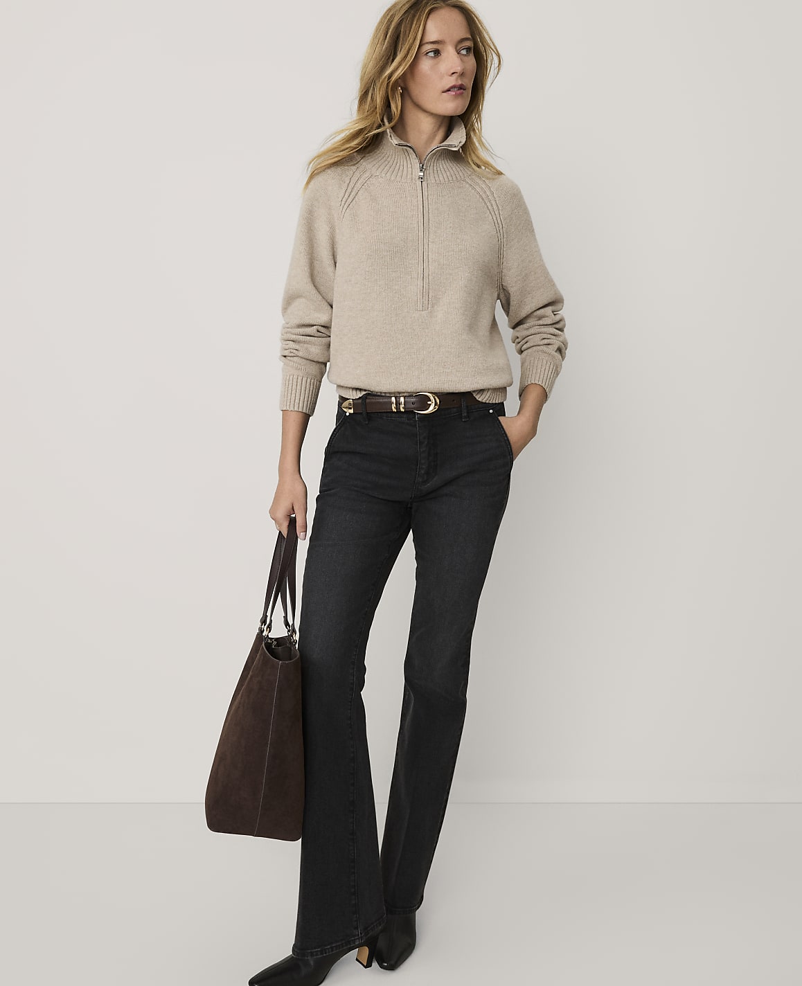 A woman wearing a beige quarter-zip sweater and dark flared jeans stands holding a brown tote bag against a plain white background.