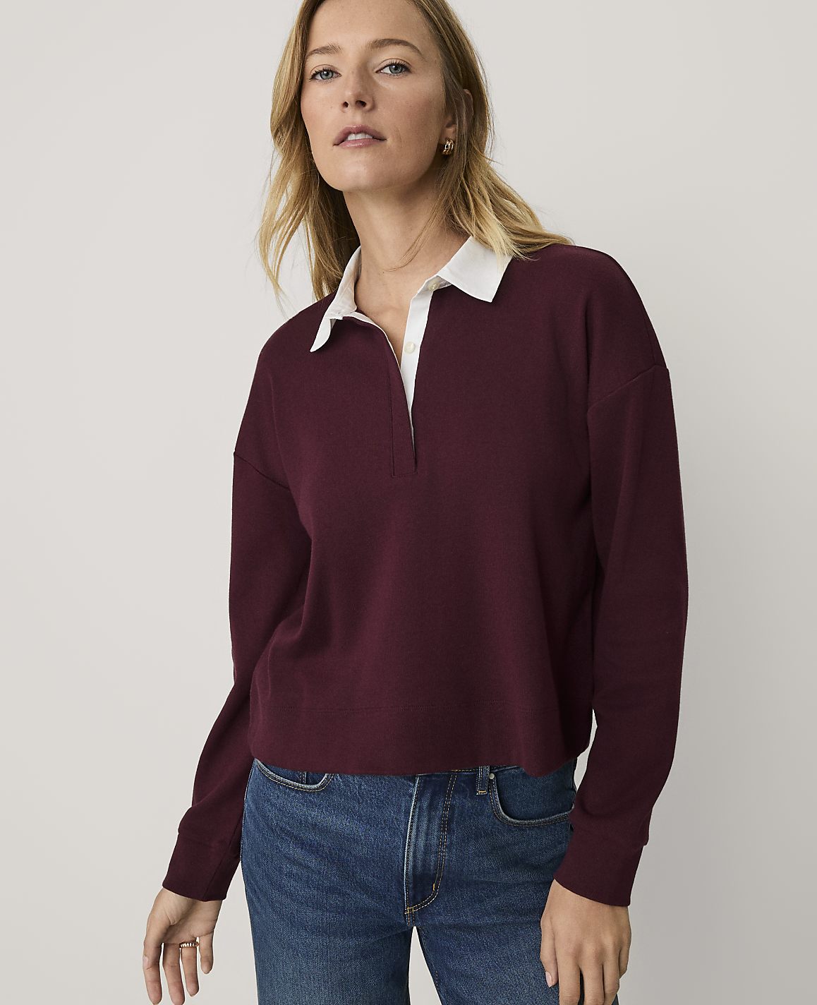 A woman wearing a maroon long-sleeved shirt with a white collar and blue jeans stands against a plain, light-colored background.
