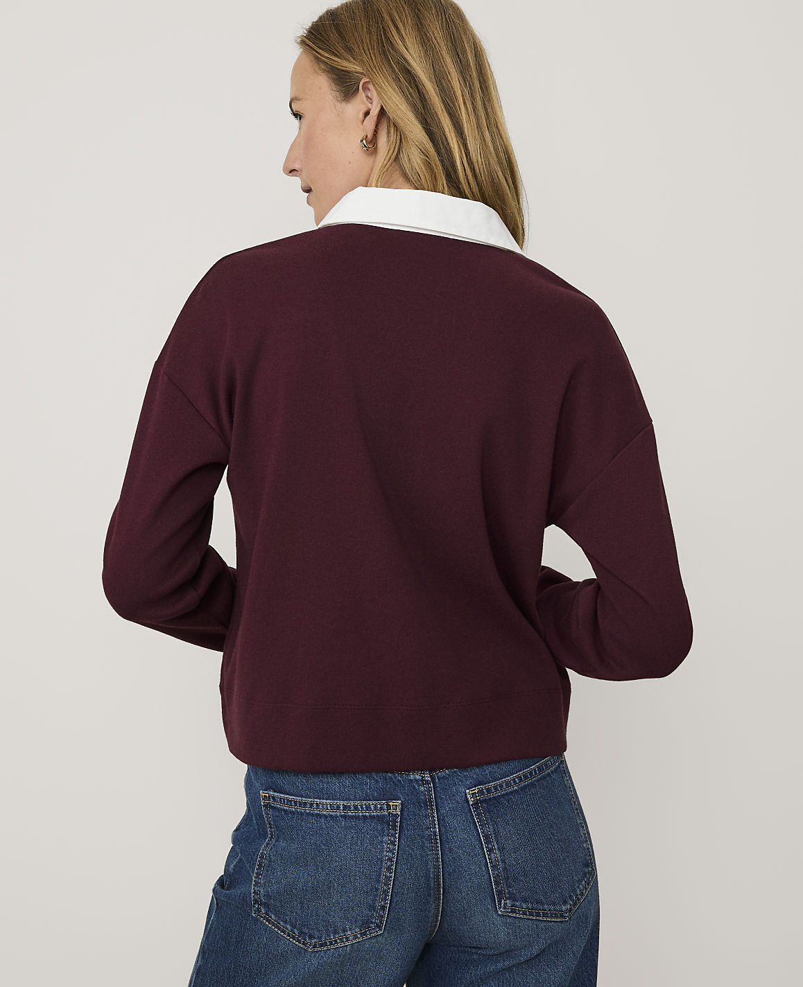 A woman wearing a maroon sweater with a white collar and blue jeans stands against a plain light gray background.
