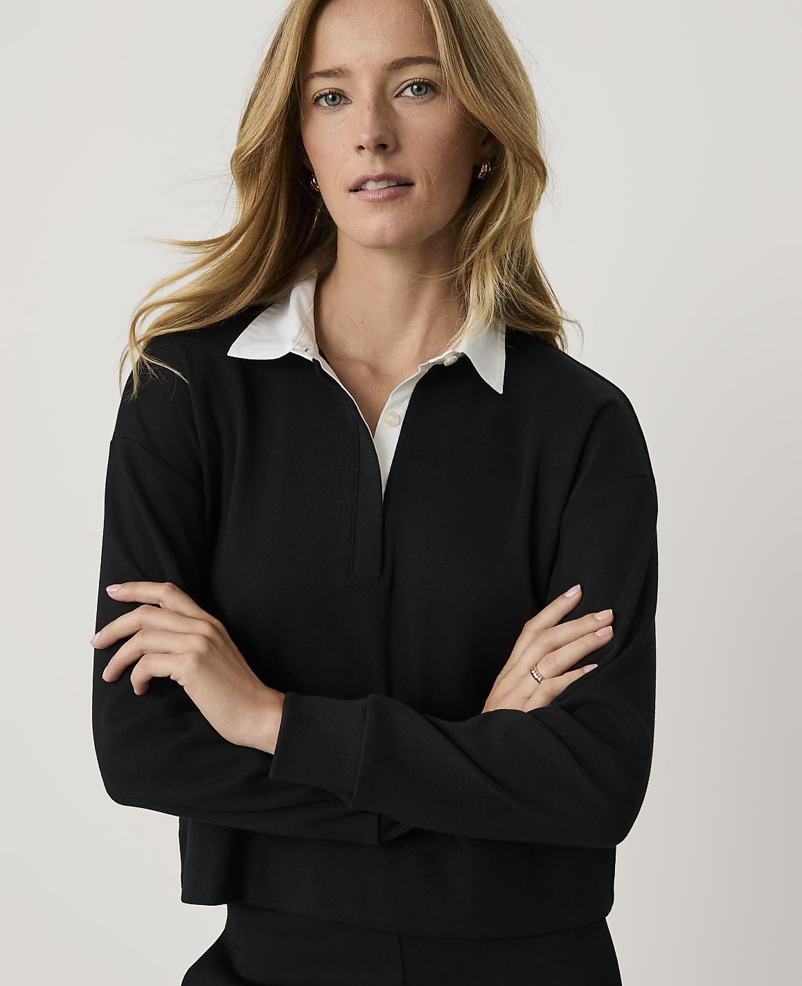 A blonde woman wearing a black long-sleeved shirt with a white collar stands with her arms crossed against a plain, light-colored background.