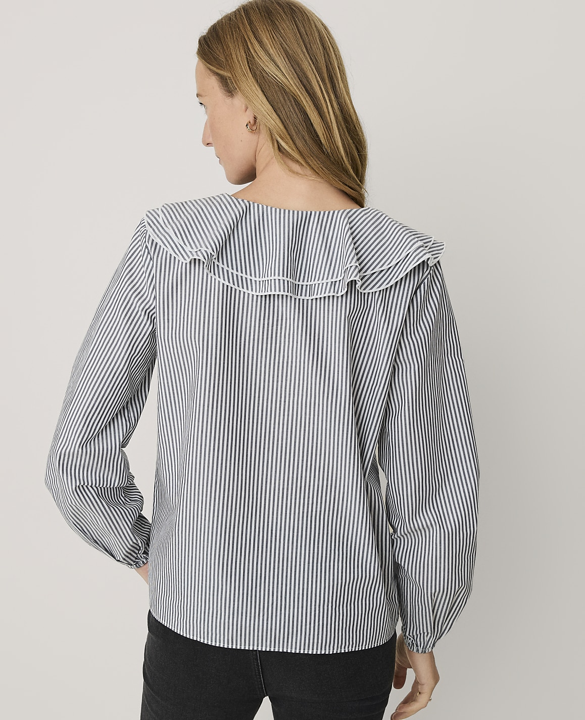 A woman wearing a blue and white striped blouse with a ruffled collar stands against a plain light gray background.