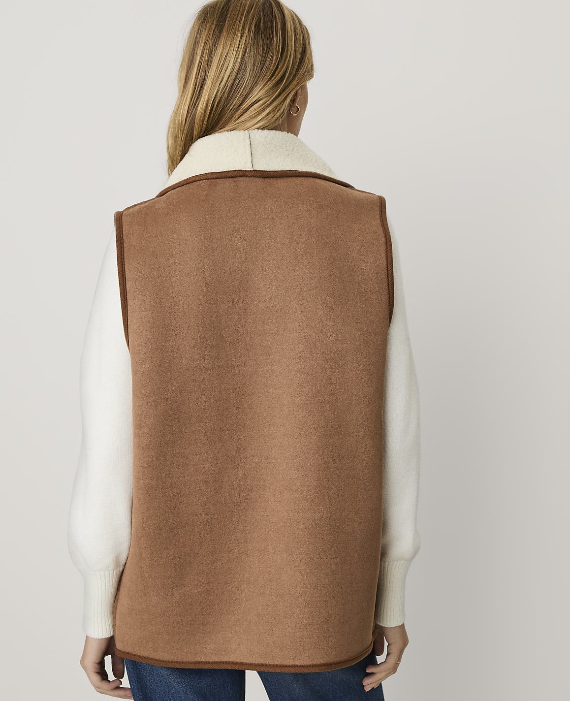 A woman viewed from behind wears a brown vest over a white sweater against a plain light gray background.