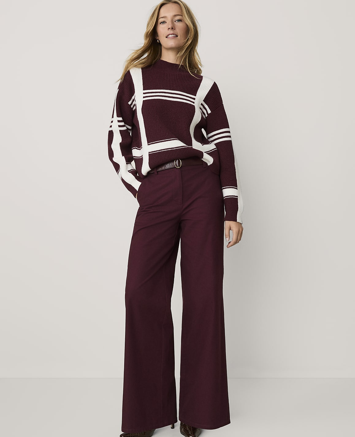 A woman wearing a maroon and white striped sweater and matching wide-leg trousers stands against a plain white background.