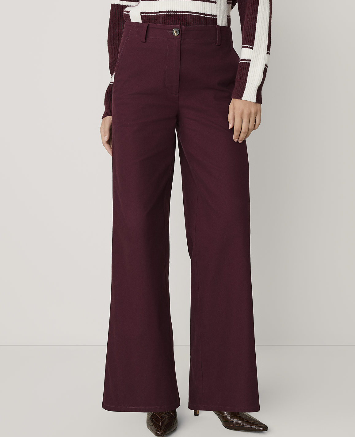 A person wears wide-leg burgundy trousers and a striped sweater against a plain, light-colored background.