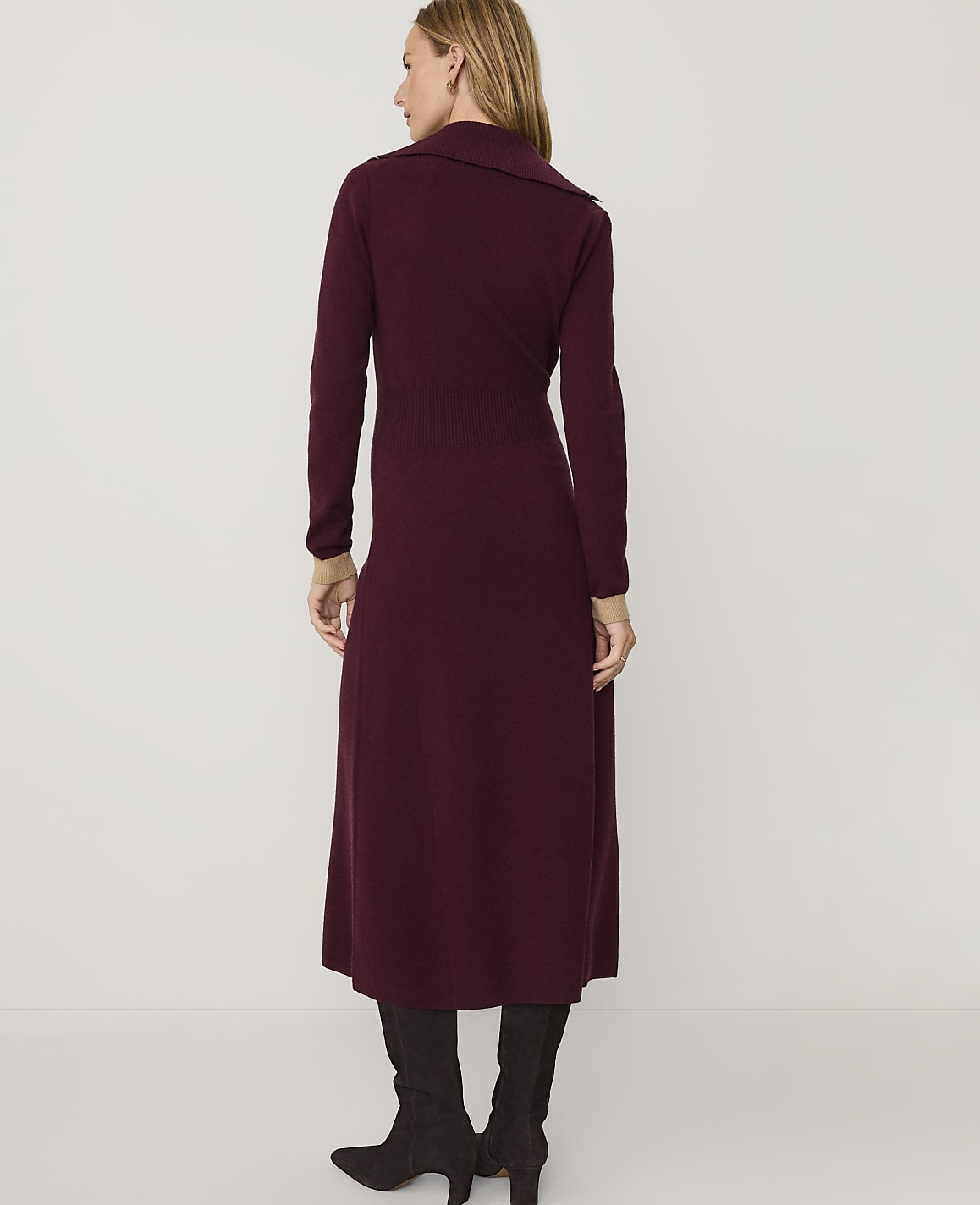 A woman wearing a long, burgundy knit dress and dark grey boots stands against a plain white background.