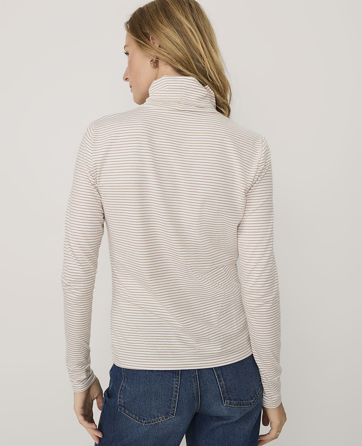 A woman wearing a striped turtleneck and blue jeans stands against a plain white background.