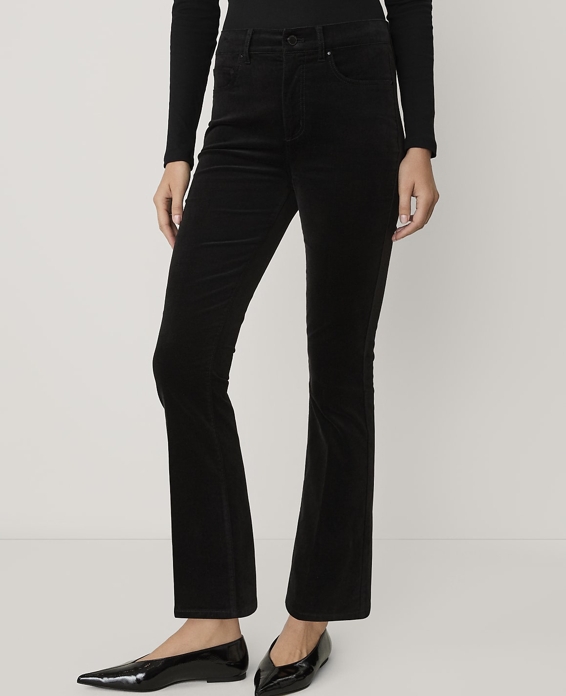A person wearing a black long-sleeved shirt, black flared corduroy trousers, and black pointed-toe flats stands against a plain white background.