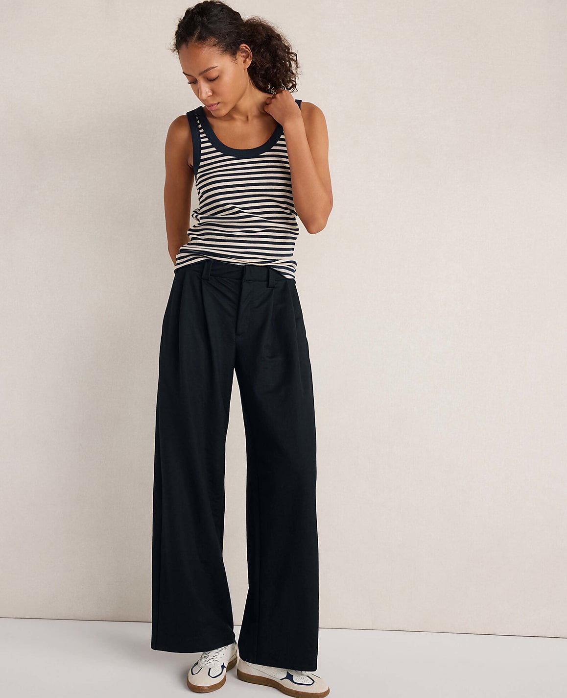 A woman wearing a striped tank top and wide-leg black trousers stands against a plain, neutral-colored wall.