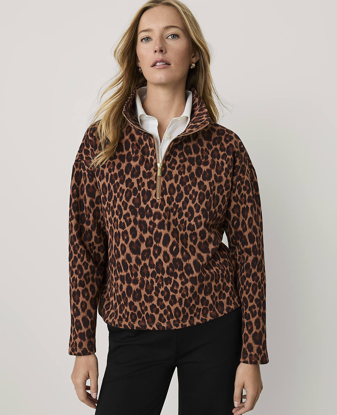 A woman wearing a leopard-print quarter-zip pullover and black pants stands against a plain, light-colored background.
