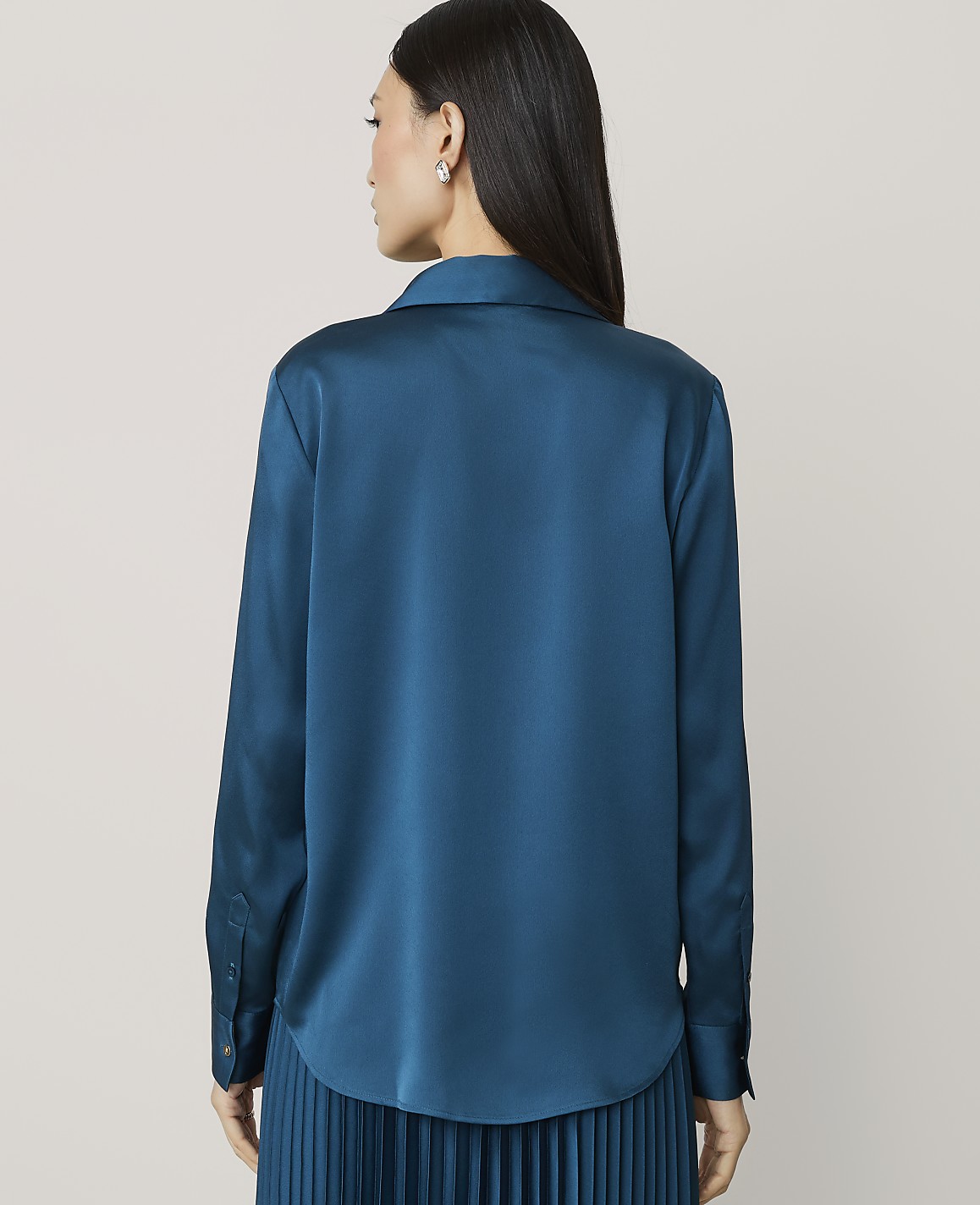 A woman wearing a teal satin shirt and pleated skirt stands with her back to the camera against a plain light gray background.
