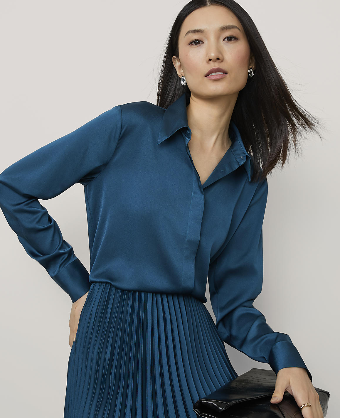 A woman wearing a teal satin shirt and pleated skirt stands against a plain, light-colored background.