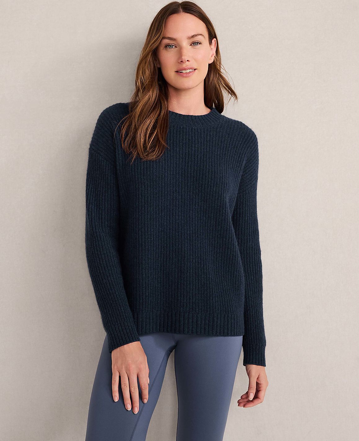 A woman in a light gray ribbed sweater stands against a textured, neutral background.
