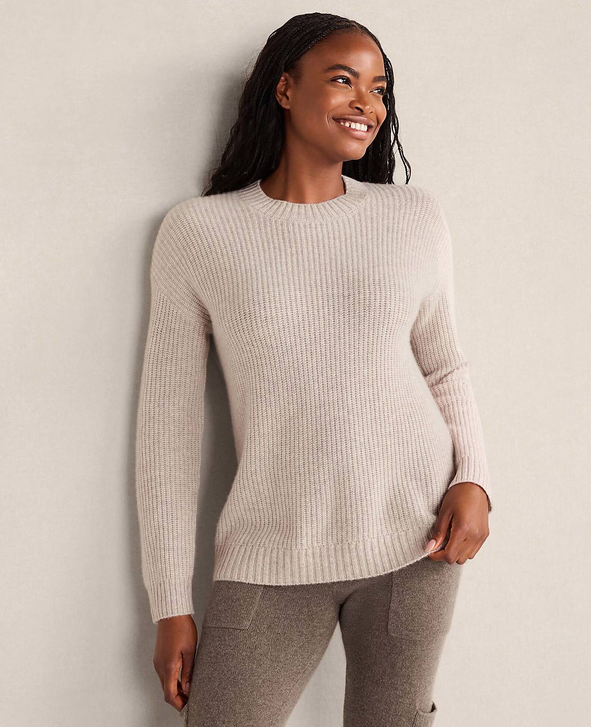 A smiling woman wearing a light beige ribbed sweater and brown pants leans against a plain, neutral-colored wall.