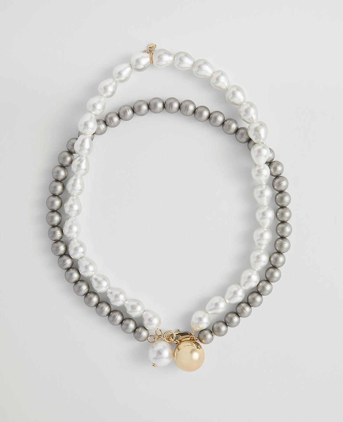 Two strands of pearls, one white and baroque and the other grey and round, are laid out in an oval shape against a plain white background.
