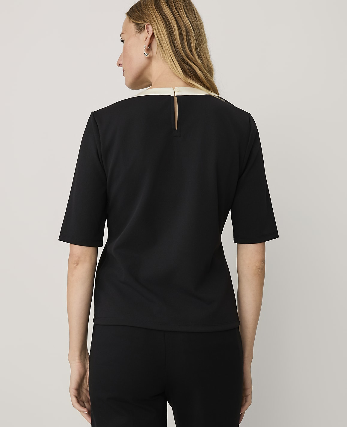 A woman wearing a black short-sleeved top and black pants stands with her back to the camera against a plain light gray background.