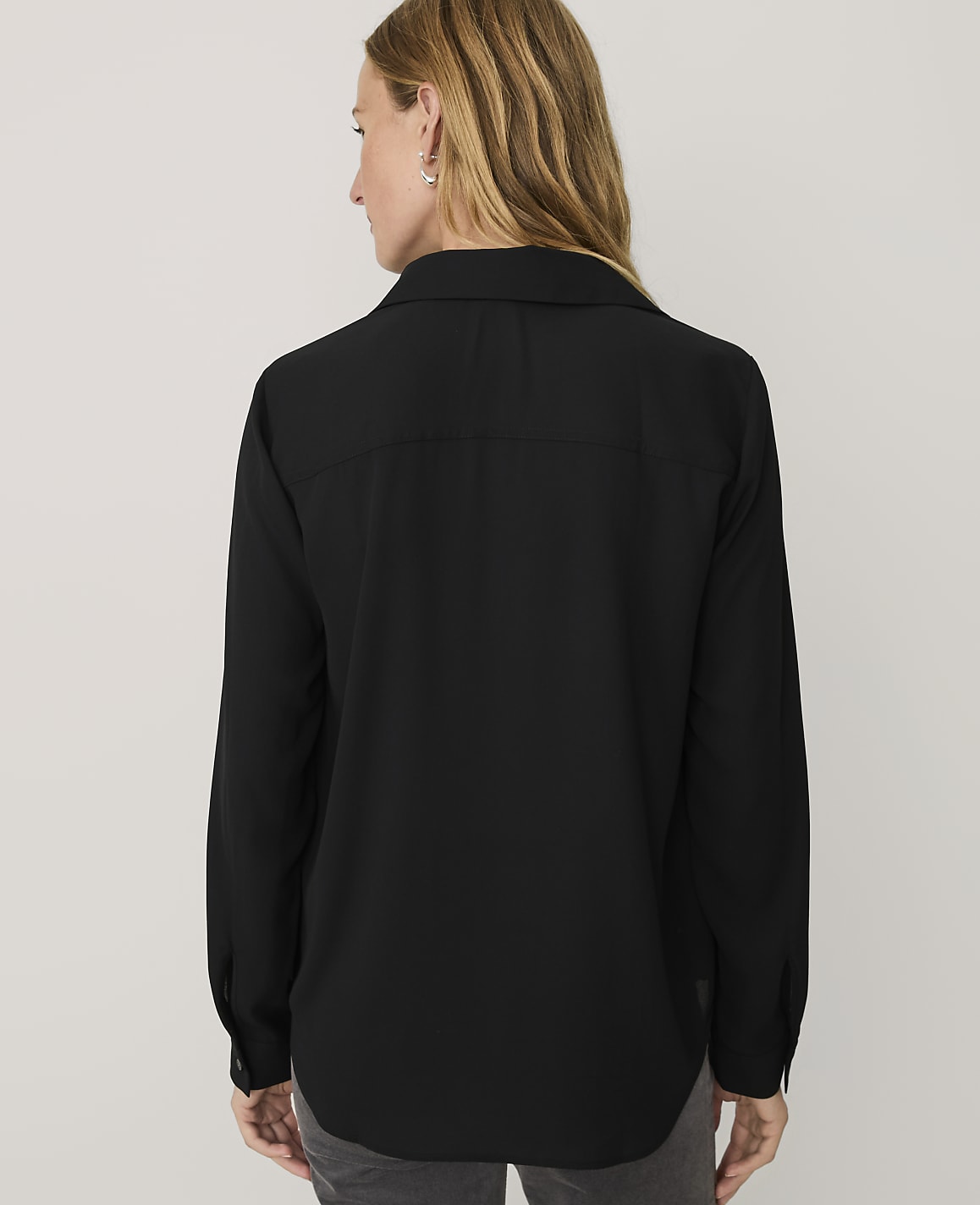 A woman wearing a black long-sleeved button-down shirt stands with her back to the camera against a plain light gray background.